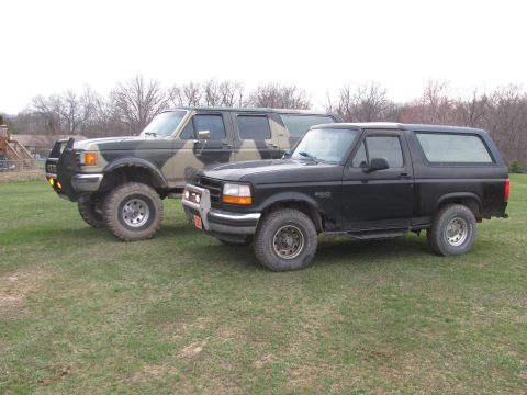 1996 Ford Bronco - A black 1996 Ford Bronco is parked next to a camouflage SUV on a grassy field.