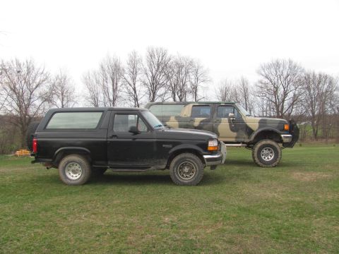 1996 Ford Bronco - Two Ford SUVs, a black Bronco and a camouflage F-Series truck, are parked side-by-side on a grassy field.