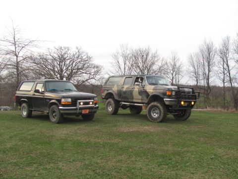 1996 Ford Bronco - Two Ford Broncos, a black 1992-1996 model and a camouflage-painted later model, sit on a grassy field under a cloudy sky.