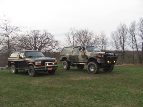 1996 Ford Bronco - Two Ford Broncos, a black model and a camouflage-painted one, are parked side-by-side on a grassy field with trees in the background.