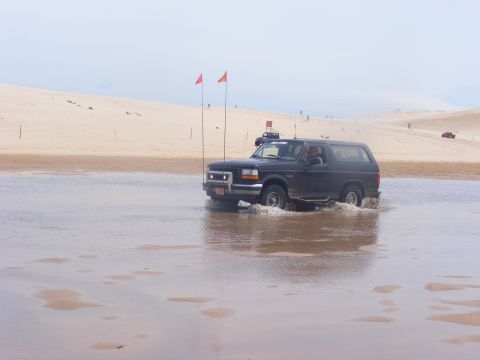 1996 Ford Bronco - A 1996 Ford Bronco navigates through a shallow body of water with sand dunes visible in the distance.