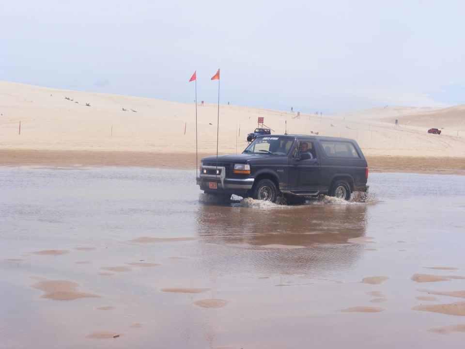Black 1996 Ford Bronco driving through shallow water with sand dunes in the background