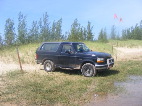1996 Ford Bronco - A black 1996 Ford Bronco is parked on a sandy area with grass and trees under a blue sky.