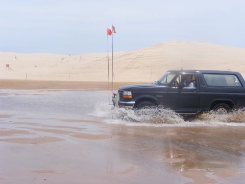 1996 Ford Bronco - A dark 1996 Ford Bronco splashes through shallow water on a sandy beach with dunes in the background.