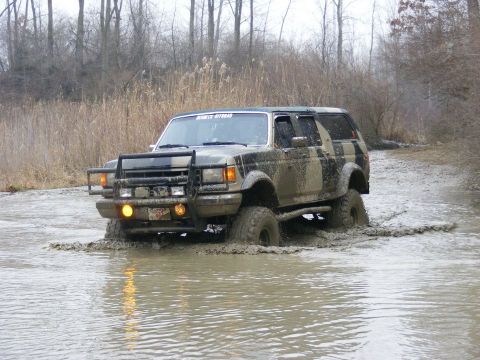 1996 Ford Bronco - This SUV is navigating through a muddy water crossing with trees and reeds in the background.