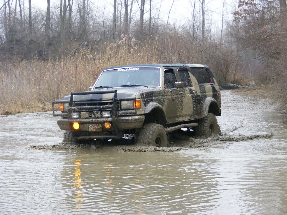 Muddy SUV driving through water, has a brush guard and camouflage paint