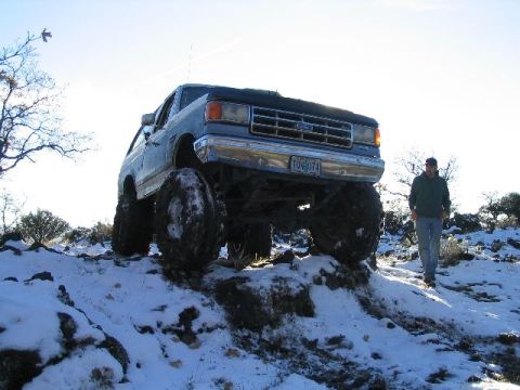 1988 Ford Bronco - A man stands next to a heavily modified Ford truck with large tires, navigating a snowy, rocky incline.