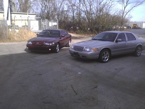 2003 Ford Crown Victoria - Two sedans, a silver Ford Crown Victoria and a maroon Mercury Grand Marquis, are parked outside.