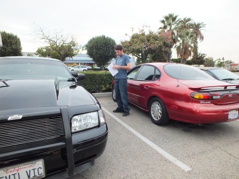 2003 Ford Crown Victoria - Black Crown Victoria nose with EVIL VIC plate parked next to a red Ford Taurus, man holding paperwork between them