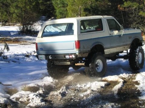 1988 Ford Bronco - Modified Ford Bronco with large tires driving on a snowy trail, kicking up snow and dirt.