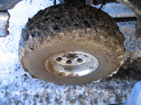 1988 Ford Bronco - A close-up view of a heavily mud-covered tire and wheel, set against a snowy backdrop.