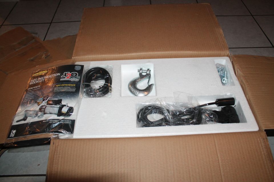 Winch and accessories in an open cardboard box. Includes instruction booklet, hook, cables, and hardware