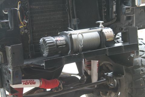 1990 Ford Bronco - A winch is mounted to the front of a vehicle, positioned in front of the radiator and grille assembly.