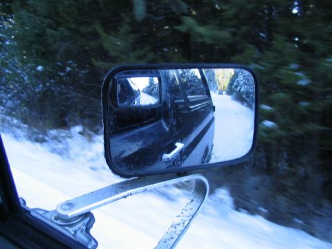 1988 Ford Bronco - Driving on a snowy road, as seen through the side mirror, with another vehicle ahead.