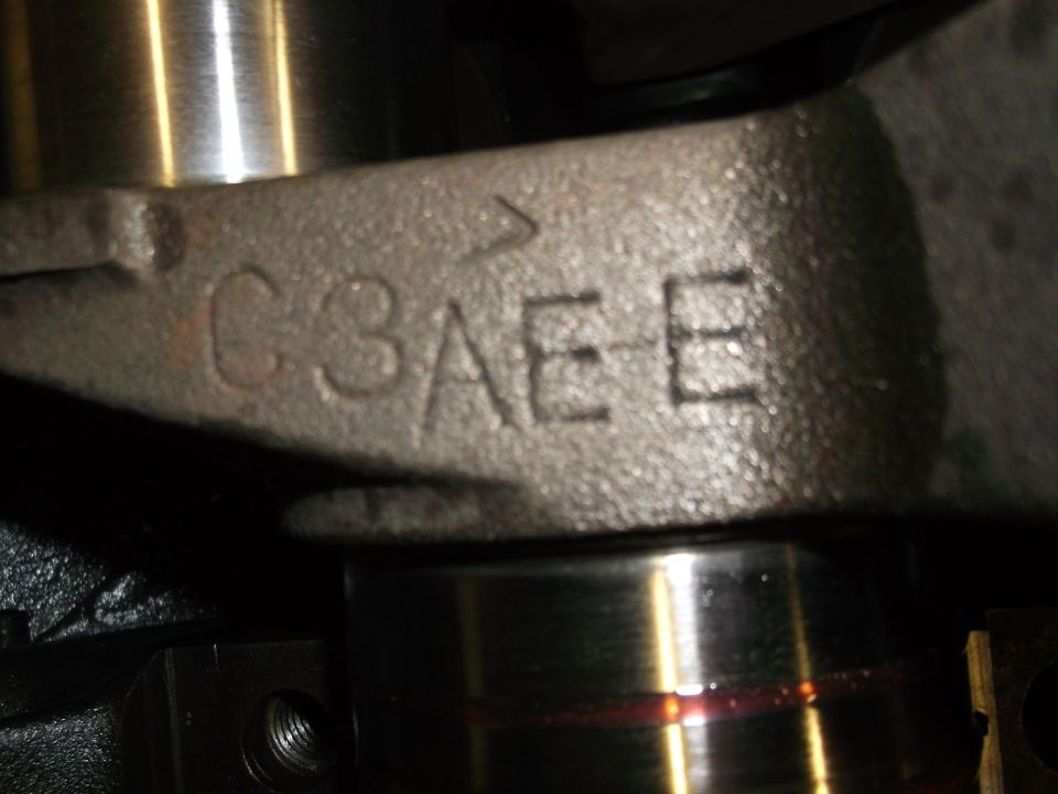 Close-up of a metal engine part with casting marks reading C3AE. A red fluid is visible