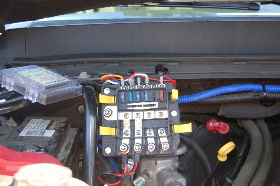 Engine bay fuse box with yellow clips, fuses, and wiring. A clear plastic cover with text sits to the left