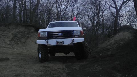 1989 Ford Bronco - A white Ford pickup truck with a custom diamond plate bumper and 4x4 badging is shown from the front on a dirt path.