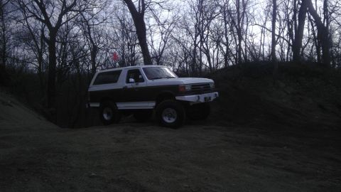 1989 Ford Bronco - A two-tone Ford Bronco is parked on a dirt hill surrounded by bare trees.