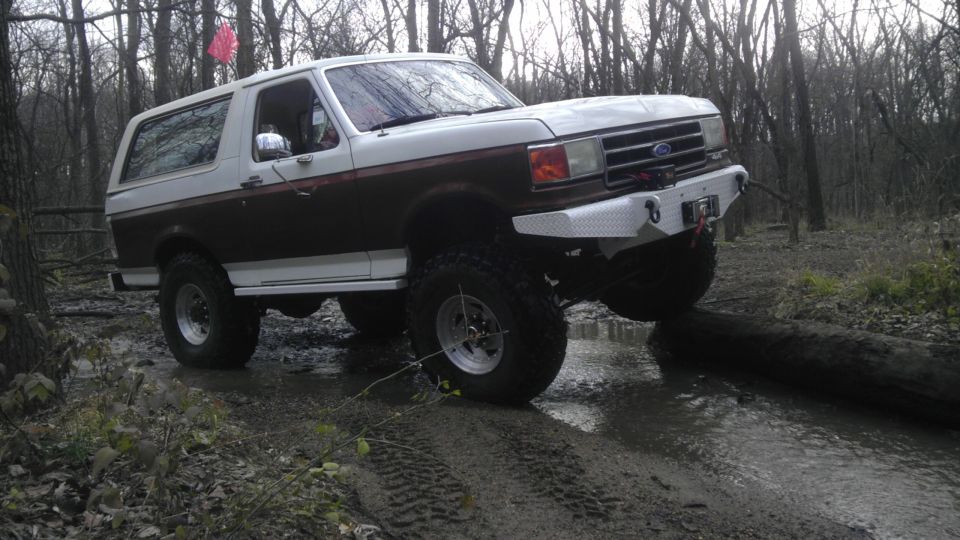 White and brown lifted 1989 Ford Bronco driving over a log in a muddy forest