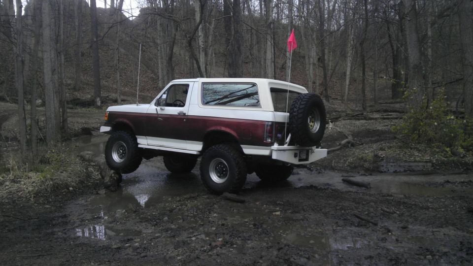 1989 Ford Bronco with lifted suspension driving through a muddy trail in a forest