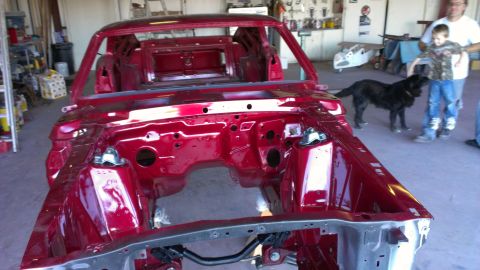 1970 Ford Mustang - A bare 1970 Ford Mustang chassis gleams red in a workshop, awaiting restoration. A dog and two people are visible in the background.