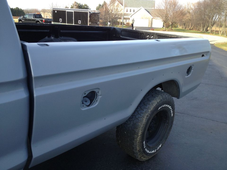 Side view of a gray pickup truck bed with a fuel door and a tire. Another truck and trailer are in the background