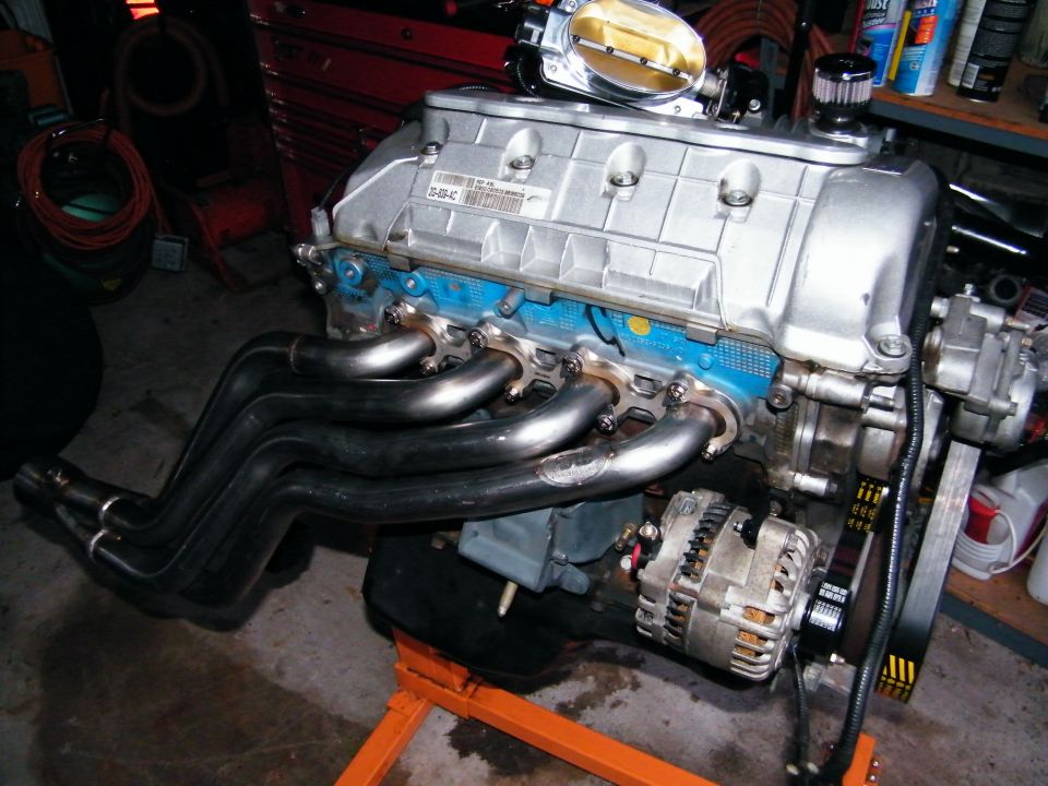 V8 engine block with blue intake manifold, chrome headers, and alternator. Part number visible on intake