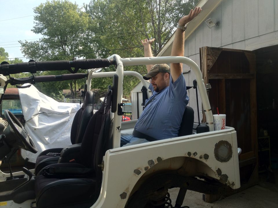 Man with arms raised sitting in a white Jeep with black seats. Some rust spots on the rear fender