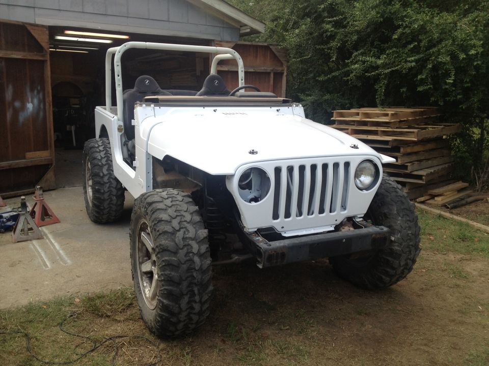 White Jeep TJ with aftermarket wheels and large off-road tires, parked in front of an open garage. Roll cage is visible