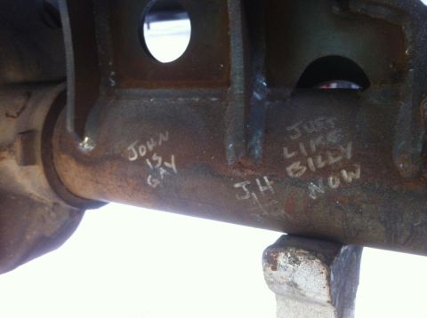 2000 Jeep TJ - Graffiti on a rusty metal part reads 'JOHN IS GAY' and 'JUST LIKE BILLY NOW'.