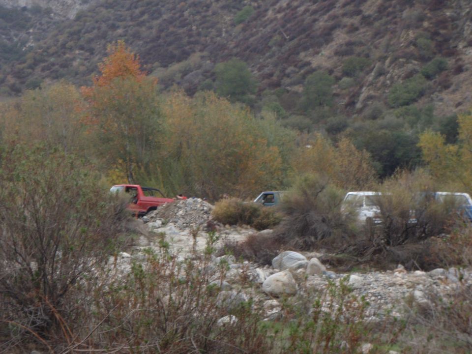 Red pickup truck and two white vans parked in a rocky, brush-filled area with hills in the background
