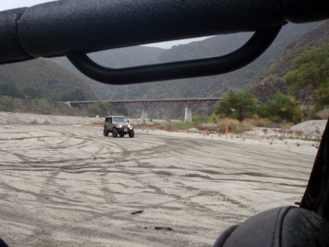 1978 Ford Bronco - A Jeep Wrangler navigates a dry riverbed, kicking up dust under a cloudy sky.
