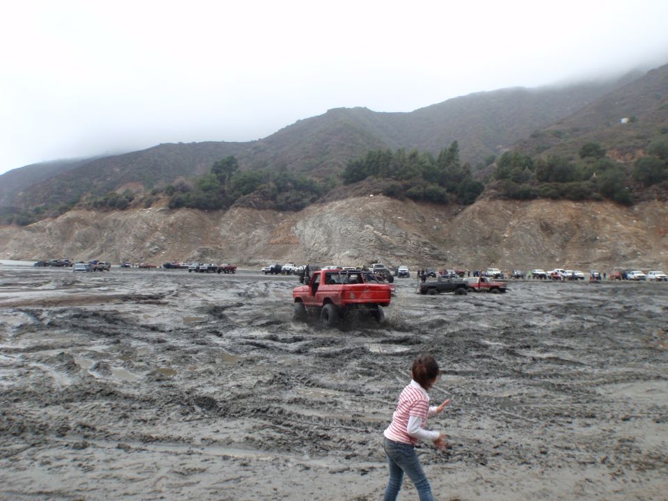 Red pickup truck driving through mud, rear view. Other vehicles and a person are visible in the muddy landscape