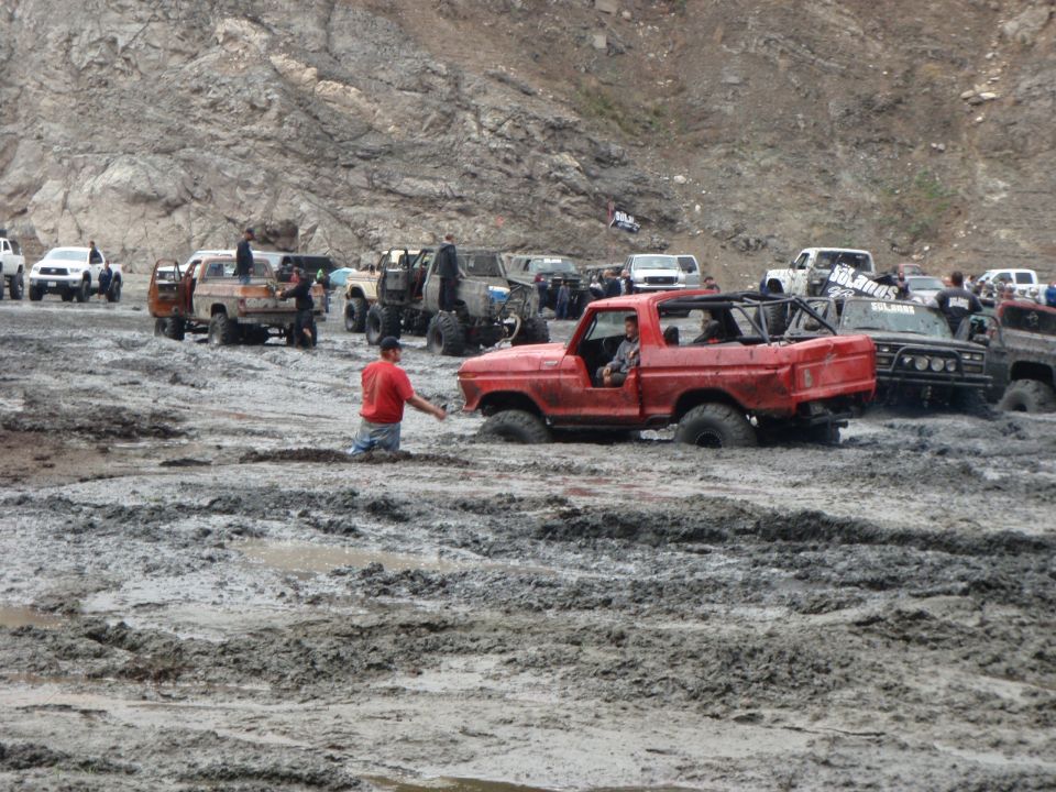 Red pickup truck stuck in mud, people around other off-road vehicles in a muddy area