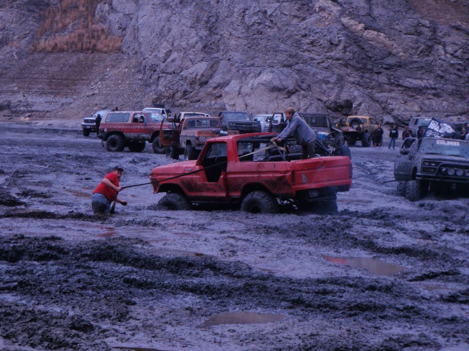 1978 Ford Bronco stuck in mud, people pulling with rope. Other trucks in background