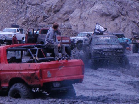 1978 Ford Bronco - Multiple off-road vehicles navigate a muddy terrain, with a red Bronco in the foreground and a person observing from its rear.