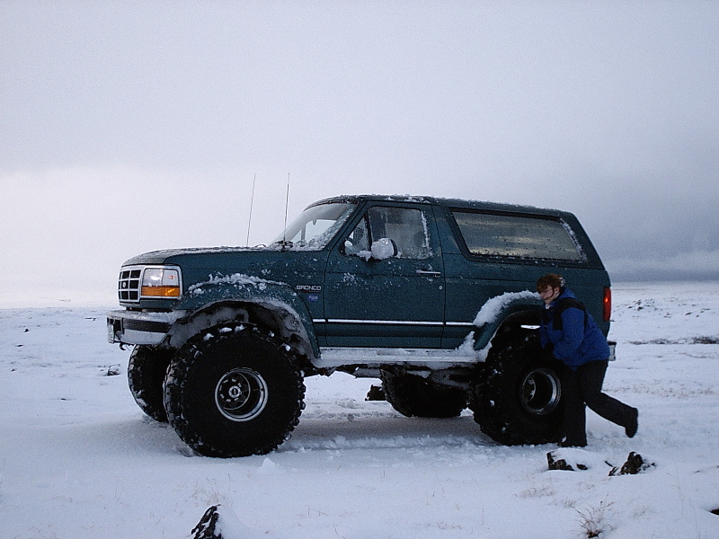 Lifted teal Ford Bronco on big tires in snow, person in blue jacket at rear