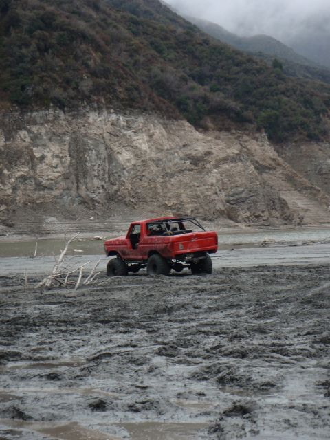 1978 Ford Bronco - A red truck navigates a muddy terrain with a steep, vegetated hillside in the background.