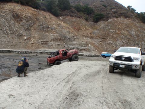 1978 Ford Bronco - A red off-road truck is stuck in the mud while a person watches and takes photos, with other vehicles nearby.