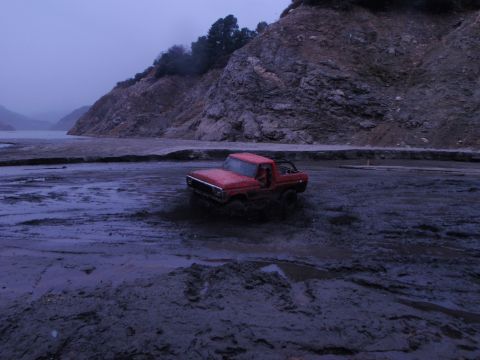 1978 Ford Bronco - Red 1978 Ford Bronco navigating through a muddy terrain with a rocky cliff in the background.
