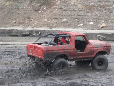 1978 Ford Bronco - Red 1978 Ford Bronco with a roll cage navigates a muddy terrain, kicking up spray.