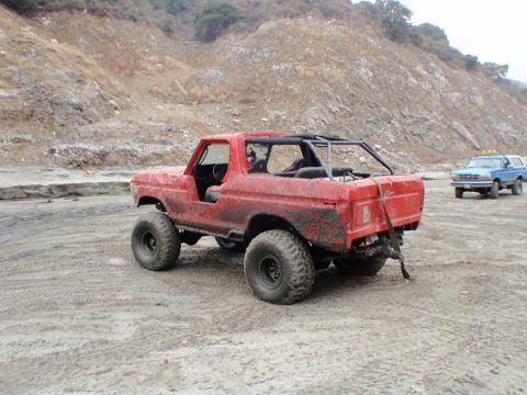 1978 Ford Bronco - A red off-road SUV with a roll cage and large tires is parked in a dirt lot, with another blue SUV in the distance.