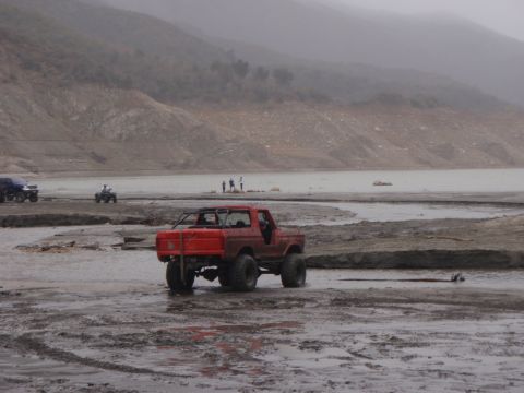 1978 Ford Bronco - A red Bronco navigates a muddy terrain near a body of water under an overcast sky.