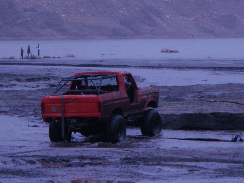 Red 1978 Ford Bronco with roll cage, mud-splattered, on a muddy shore