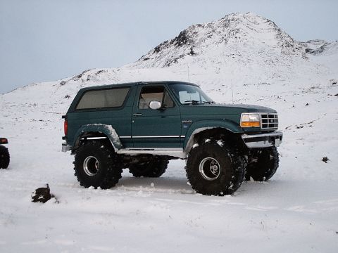1996 Ford Bronco - A green 1996 Ford Bronco equipped with oversized tires navigates a snowy terrain, with a snow-covered mountain in the background.