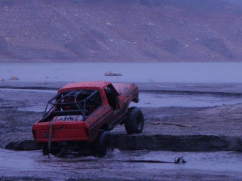 1978 Ford Bronco - A red off-road vehicle with a prominent roll cage navigates through a muddy terrain under a dusky sky.