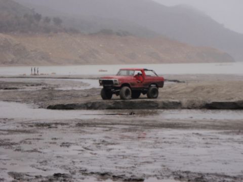 1978 Ford Bronco - A red 1978 Ford Bronco navigates a muddy, off-road terrain with hills in the background.