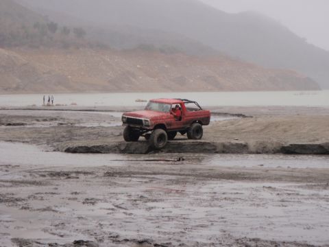 1978 Ford Bronco - A red 1978 Ford Bronco navigates a muddy terrain near a body of water, with hills in the background.