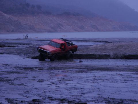 1978 Ford Bronco - A red off-road vehicle is stuck in the mud near a body of water, with a few people visible in the distance.