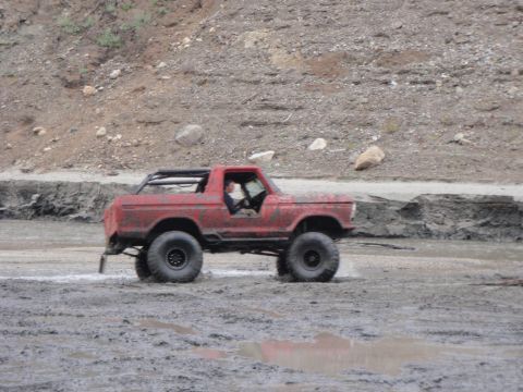 1978 Ford Bronco - A red 1978 Ford Bronco navigates a muddy terrain, its large off-road tires kicking up spray.